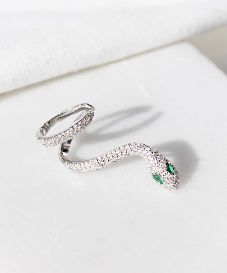 May include: A silver-toned snake ring with a wrap-around design. The ring is encrusted with small, clear crystals along the body, with two green gems for eyes. The ring is set against a white background.