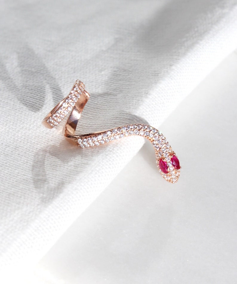 May include: Rose gold snake ring with clear and red gemstones. The ring wraps around the finger in a serpentine design, with the head featuring two red gemstones for eyes. The ring is set against a white background.