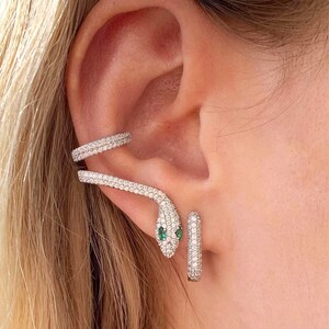May include: A silver snake ear cuff and matching hoop earrings. The snake ear cuff wraps around the ear, with a head featuring two green eyes. Both pieces are encrusted with small, sparkling stones.
