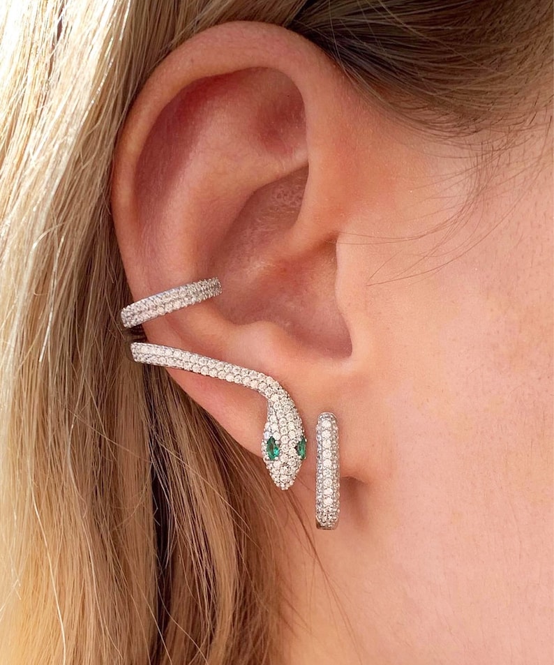 May include: A silver snake ear cuff and matching hoop earrings. The snake ear cuff wraps around the ear, with a head featuring two green eyes. Both pieces are encrusted with small, sparkling stones.