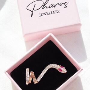 May include: A rose gold snake ring with clear and red gemstones, presented in a pink box with the text "Pharos JEWELLERY". The ring is coiled and detailed, set against a black velvet interior.