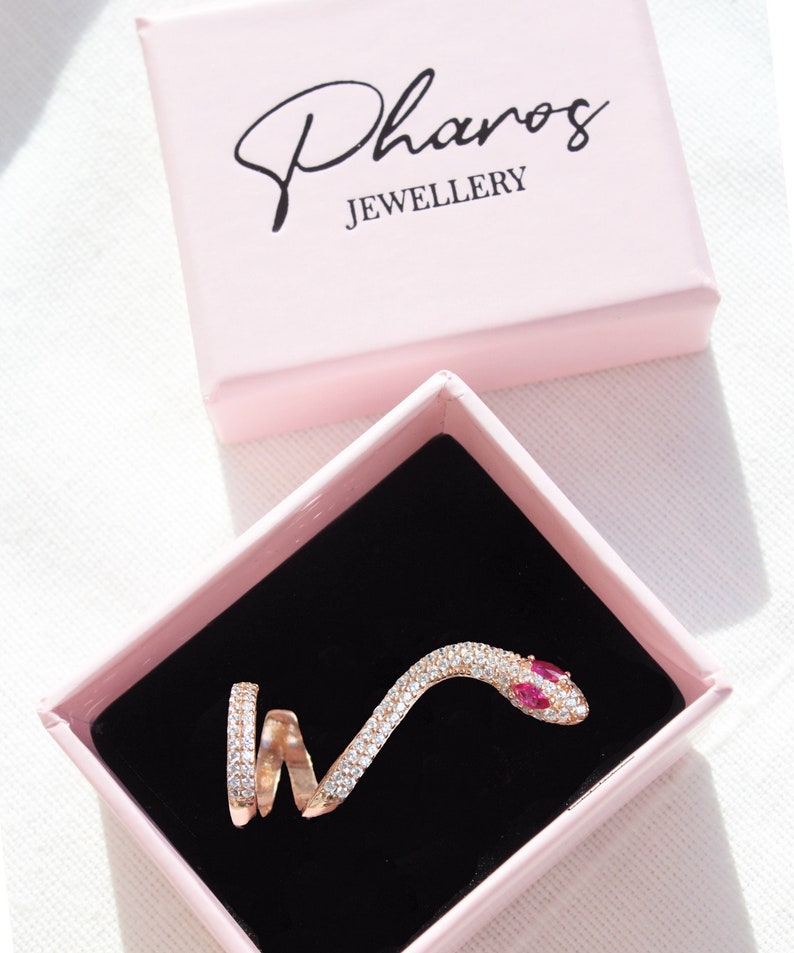 May include: A rose gold snake ring with clear and red gemstones, presented in a pink box with the text "Pharos JEWELLERY". The ring is coiled and detailed, set against a black velvet interior.
