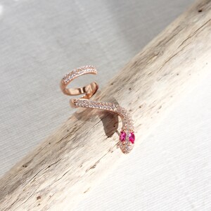 May include: Rose gold snake ring with a wrap-around design. The ring features a head with two pink gemstones for eyes and is encrusted with small, clear gemstones along its body. The ring is displayed on a piece of driftwood.