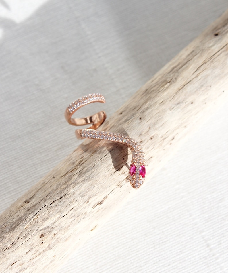 May include: Rose gold snake ring with a wrap-around design. The ring features a head with two pink gemstones for eyes and is encrusted with small, clear gemstones along its body. The ring is displayed on a piece of driftwood.