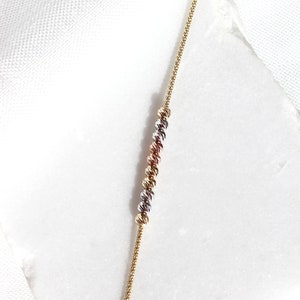 Puede incluir: Un delicado collar de oro con una serie de cuentas esf&eacute;ricas texturizadas en oro, plata y oro rosa. La fina cadena y las cuentas espaciadas uniformemente crean un dise&ntilde;o sutil pero elegante. El collar se muestra sobre una superficie blanca.