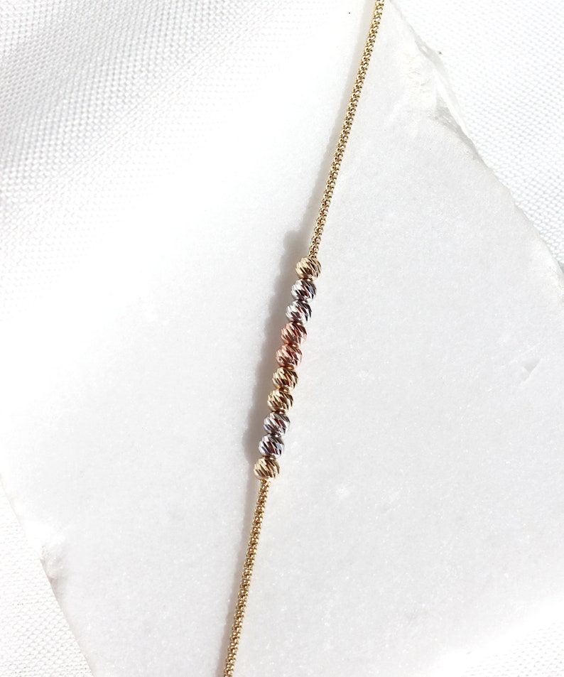 Puede incluir: Un delicado collar de oro con una serie de cuentas esf&eacute;ricas texturizadas en oro, plata y oro rosa. La fina cadena y las cuentas espaciadas uniformemente crean un dise&ntilde;o sutil pero elegante. El collar se muestra sobre una superficie blanca.