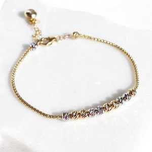 Puede incluir: Pulsera dorada con una cadena delicada y una serie de cuentas texturizadas en oro, plata y oro rosa. La pulsera ajustable se muestra sobre una superficie blanca, mostrando su elegante dise&ntilde;o.
