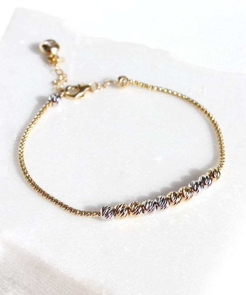 Puede incluir: Pulsera dorada con una cadena delicada y una serie de cuentas texturizadas en oro, plata y oro rosa. La pulsera ajustable se muestra sobre una superficie blanca, mostrando su elegante dise&ntilde;o.