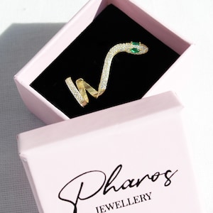 May include: A gold-colored snake ring with clear and green gemstones, displayed in a pink jewelry box with the brand name "Pharos JEWELLERY" in black script.