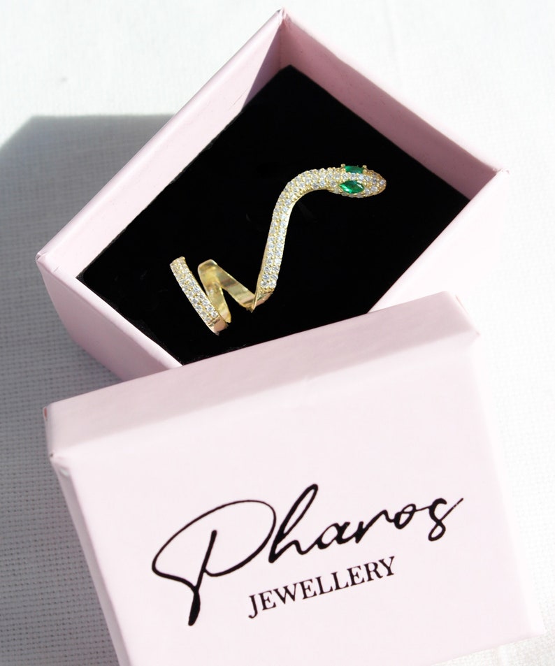 May include: A gold-colored snake ring with clear and green gemstones, displayed in a pink jewelry box with the brand name "Pharos JEWELLERY" in black script.
