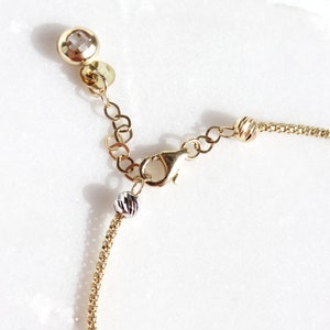 Puede incluir: Primer plano de un collar dorado con cierre de mosquet&oacute;n y cadena ajustable. El collar presenta un peque&ntilde;o dije redondo y facetado y una peque&ntilde;a cuenta plateada. La cadena es delicada y tiene un dise&ntilde;o texturizado.
