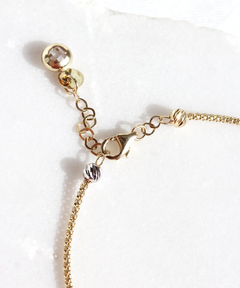 Puede incluir: Primer plano de un collar dorado con cierre de mosquet&oacute;n y cadena ajustable. El collar presenta un peque&ntilde;o dije redondo y facetado y una peque&ntilde;a cuenta plateada. La cadena es delicada y tiene un dise&ntilde;o texturizado.