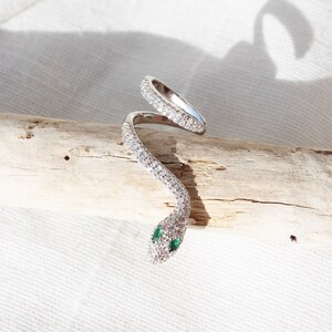 May include: A silver-toned snake ring with a wrap-around design. The ring is encrusted with clear, sparkling stones, and the snake's eyes are small green stones. The ring is displayed on a piece of driftwood against a white background.