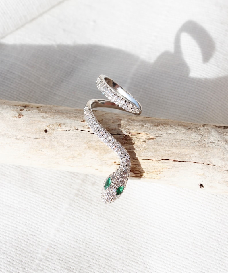 May include: A silver-toned snake ring with a wrap-around design. The ring is encrusted with clear, sparkling stones, and the snake's eyes are small green stones. The ring is displayed on a piece of driftwood against a white background.