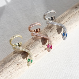 May include: Four snake-shaped ear cuffs in gold, rose gold, and silver tones. Each cuff is encrusted with small, clear crystals. The snake heads feature green, pink, and blue gemstone eyes. The jewelry is displayed on a piece of driftwood.