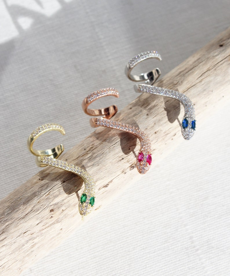 May include: Four snake-shaped ear cuffs in gold, rose gold, and silver tones. Each cuff is encrusted with small, clear crystals. The snake heads feature green, pink, and blue gemstone eyes. The jewelry is displayed on a piece of driftwood.