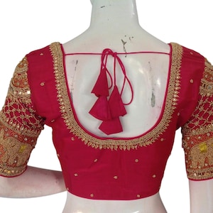 Puede incluir: Una blusa de seda roja con bordados y adornos dorados. La blusa tiene un escote en V profundo y mangas cortas. El bordado presenta un patrón geométrico y pequeñas piedras brillantes.