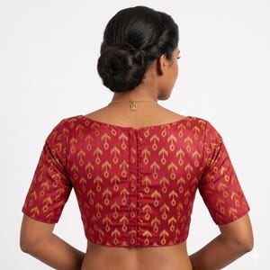 May include: A maroon blouse with a gold geometric pattern. The short-sleeved top has a boat neck and a row of small buttons down the back. The fabric has a subtle sheen.