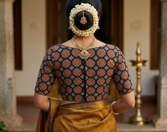 Puede incluir: Una mujer vista de espaldas, con una blusa negra con un estampado floral rojo y naranja. La blusa tiene mangas cortas y cuello barco. Lleva un collar dorado y flores en el pelo. Un sari dorado está drapeado.
