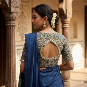 May include: A blue saree draped over a patterned blouse with a keyhole back. The blouse features a floral design in shades of green and blue, with small mirror accents. The woman is wearing gold earrings and a bracelet.
