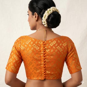 Peut inclure: Un chemisier orange avec un motif géométrique doré et des manches courtes. Le chemisier a une rangée de boutons orange dans le dos et une encolure dégagée. La modèle porte une couronne de fleurs et un collier doré.