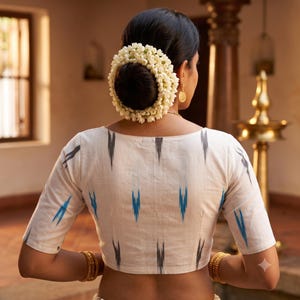 May include: A white blouse with short sleeves and a blue and grey ikat pattern. The garment features a wide neckline and is worn with a large, circular floral hair accessory made of small white blossoms arranged in a dense, rounded cluster.
