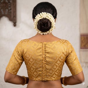 May include: A golden-yellow blouse with a subtle patterned design and short sleeves. The blouse features a row of small buttons down the back. The model's hair is styled in a bun and adorned with a ring of white flowers. Gold bangles and a necklace are also visible.