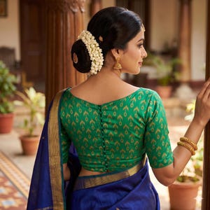 May include: A woman wearing a green blouse with a gold pattern and a blue sari. The blouse has a scoop neck and button closure down the back. The woman has her hair up in a bun adorned with white flowers and gold earrings.
