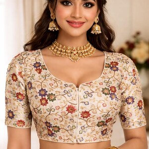 Könnte beinhalten: Cremefarbene Bluse mit kurzen Ärmeln, verziert mit farbenfroher Blumenstickerei. Die Bluse hat einen Rundhalsausschnitt und eine Knopfleiste. Das Model trägt Goldschmuck, darunter eine Halskette, Ohrringe und Armreifen.