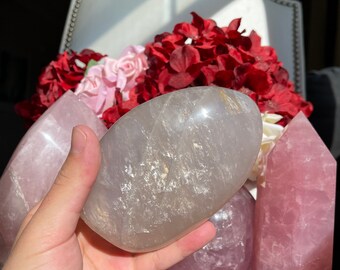 May include: A collection of rose quartz crystals in various shapes and sizes, including a large, smooth, oval-shaped crystal, a smaller sphere, and a tall, pointed crystal. The crystals are a pale pink colour and have a translucent appearance.