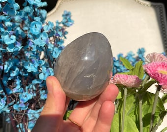 Puede incluir: Un cristal gris en forma de huevo con una superficie lisa y pulida. El cristal tiene una apariencia translúcida con sutiles estrías en todo.