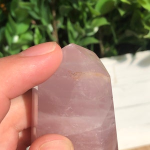 Puede incluir: Una punta de cristal de color p&uacute;rpura claro con una parte superior facetada. El cristal se sostiene en una mano, con un fondo borroso de hojas verdes. El cristal tiene una superficie lisa y pulida y un aspecto ligeramente transl&uacute;cido.