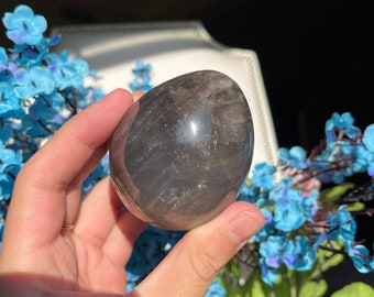 Puede incluir: Un cristal liso de forma ovalada, de color gris, con una apariencia de cuarzo ahumado. El cristal está siendo sostenido en una mano.