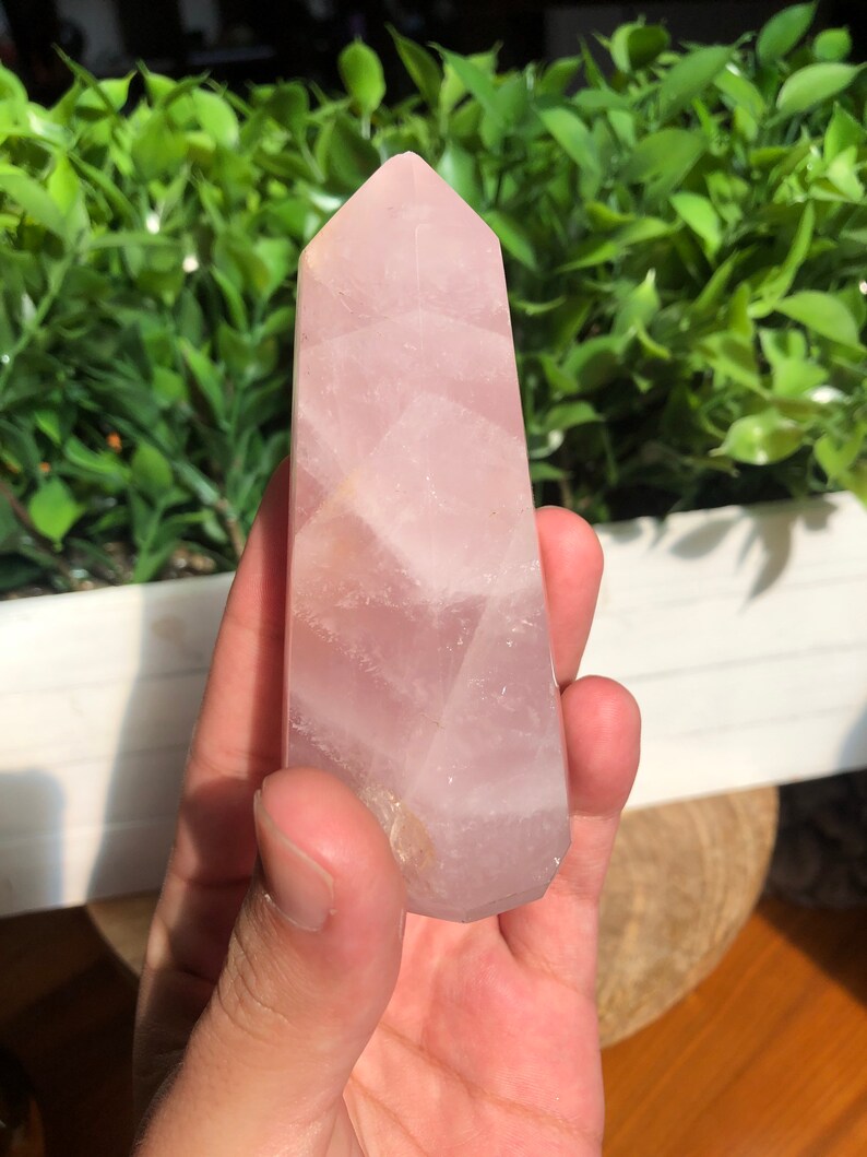 Puede incluir: Un obelisco de cuarzo rosa de color rosa claro, con un dise&ntilde;o facetado. El cristal se sostiene en una mano, con follaje verde en el fondo. El obelisco tiene una superficie pulida y una parte superior puntiaguda.