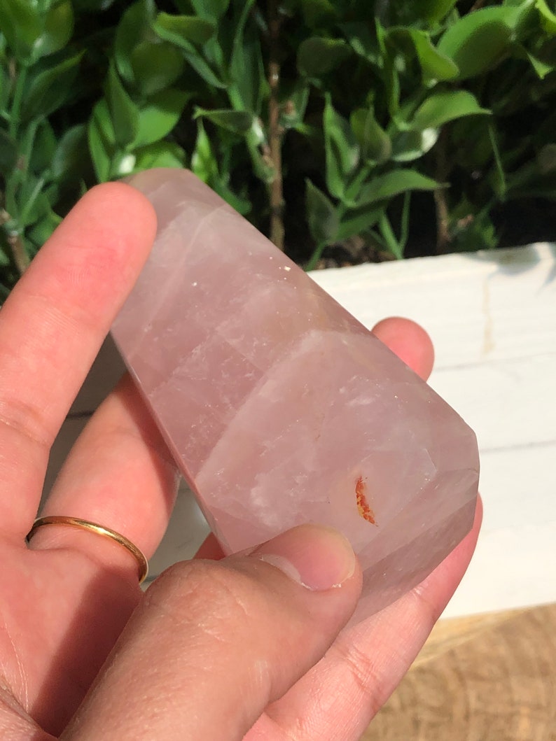 Puede incluir: Un obelisco de cristal de cuarzo rosa claro. El cristal se sostiene en una mano, y el fondo es follaje verde borroso. El obelisco tiene una superficie lisa y pulida y una forma ligeramente c&oacute;nica.