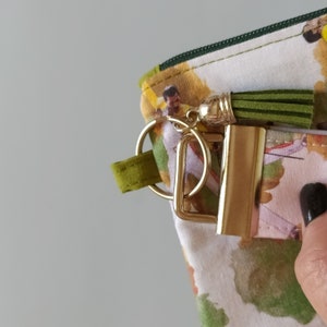 May include: Close-up of a patterned fabric pouch with a gold-colored zipper pull and a green tassel. The pouch has a gold-colored metal clasp and a green fabric loop attached to a gold ring.