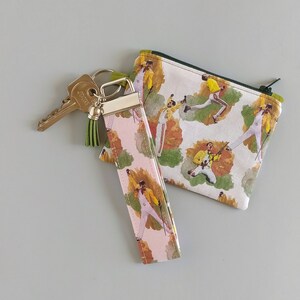 May include: A keychain, wristlet, and zippered pouch set. The items feature a repeating pattern of a singer in a yellow jacket and white pants. The wristlet and pouch have a white background with green and brown accents. The keychain has a pink background.