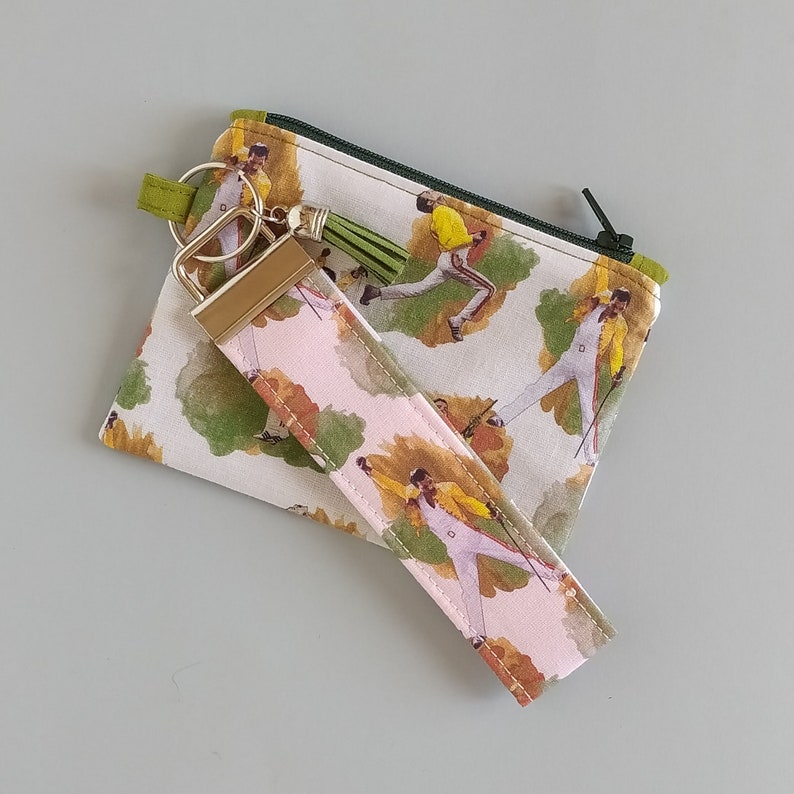 May include: A small zippered pouch, a wristlet key fob, and a keychain. The items feature a repeating pattern of a person in a white jumpsuit and yellow jacket. The pouch has a green zipper and trim. The wristlet has a gold-colored clasp and a green tassel.