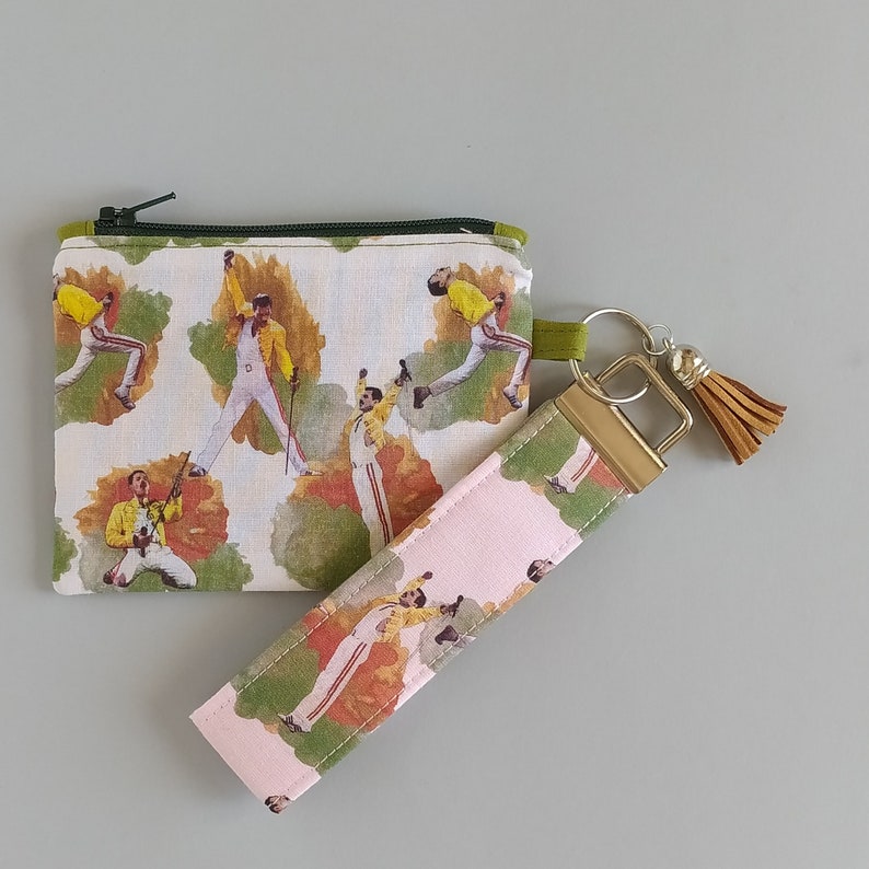 May include: A small zippered pouch and a wristlet key fob. Both items feature a repeating pattern of a performer in a yellow jacket and white pants. The pouch has a green zipper and trim. The key fob has a gold-colored metal clasp and a brown tassel.