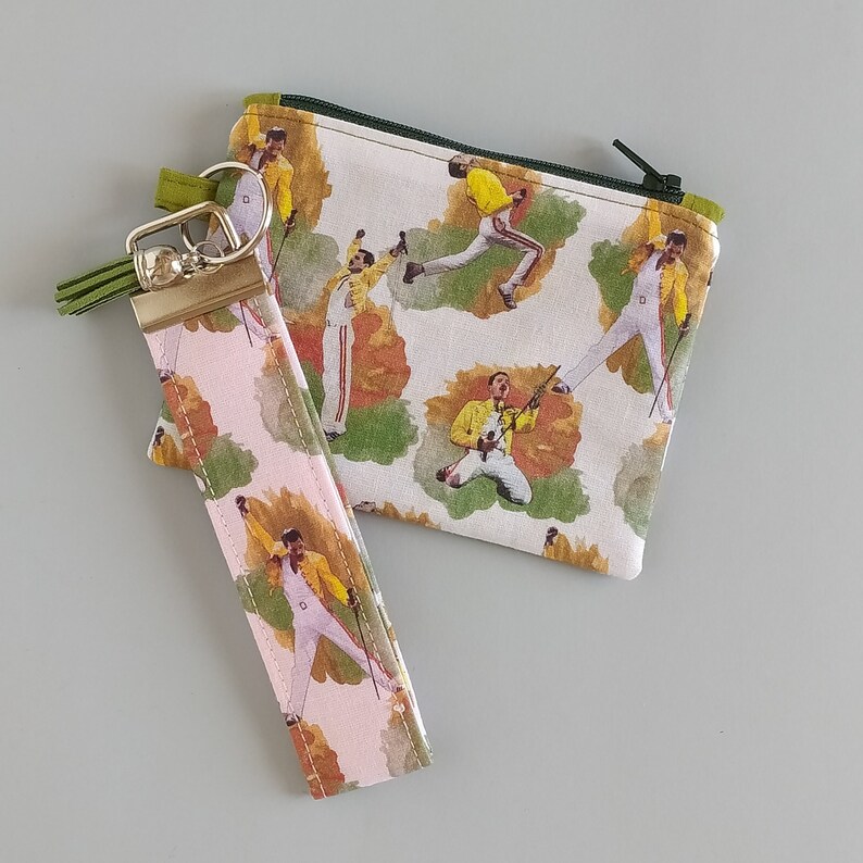 May include: A set of accessories featuring a repeating pattern of a performer in a yellow jacket and white pants. The set includes a small zippered pouch with a green zipper and a wristlet with a gold-colored clasp and green tassel.