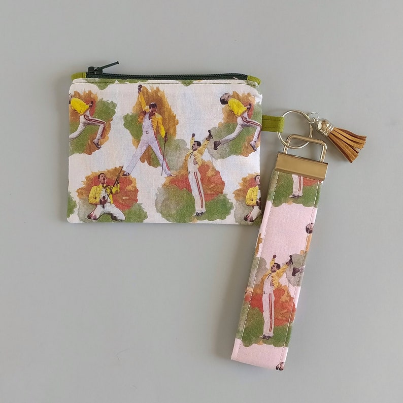 May include: A small zippered pouch and a wristlet keychain featuring a repeating pattern of a musician in a yellow jacket and white pants. The pouch has a green zipper and the keychain has a gold-colored clasp and a brown tassel.