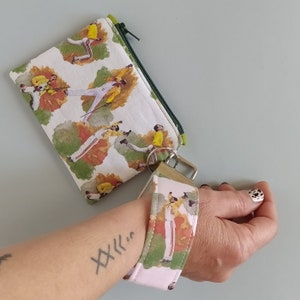 May include: A white zippered pouch and wristlet featuring a repeating pattern of a person in a yellow jacket and white pants. The pouch has a green zipper and the wristlet has a silver clasp.