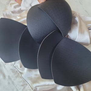 May include: Black bra cups are arranged in a fan shape, held by a hand. The cups are made of a textured material with a smooth, white edge. The background is a light-colored, silky fabric.