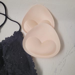 May include: Two beige bra inserts with a heart-shaped design are displayed next to a black lace bra. The inserts are triangular and have a soft, padded appearance. The bra features intricate floral lace detailing.