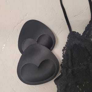May include: Black bra inserts and a black lace bralette. The bra inserts are a dark matte black, with a curved shape and a heart-shaped indentation. The bralette has delicate lace detailing and thin straps.