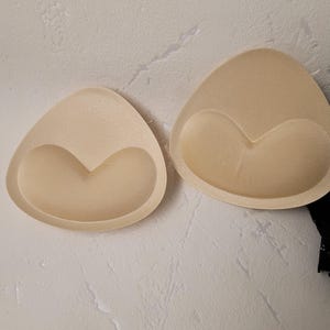 May include: Two beige, triangular bra inserts with a heart-shaped indentation. The inserts are made of a soft material and are designed to provide shape and support. The image is taken from a top-down perspective.