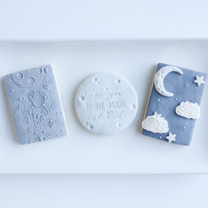 May include: A white rectangular platter holds three decorated cookies. One cookie is round with the text "love you to the moon and back." Two rectangular cookies feature an astronaut and a moon and stars design. The third cookie has a moon, stars, and clouds.