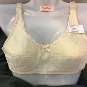 May include: Cream-coloured bra with a floral design, displayed on a mannequin. It has adjustable straps, a small bow, and a tag that says "Remove Tags Before Wearing Thank You". The bra has a delicate lace trim.