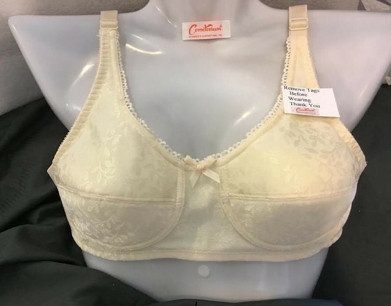 May include: Cream-coloured bra with a floral design, displayed on a mannequin. It has adjustable straps, a small bow, and a tag that says "Remove Tags Before Wearing Thank You". The bra has a delicate lace trim.