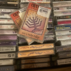 May include: A collection of vintage cassette tapes. The focus is on a cassette labeled "CHANUKAH A Singing" with a menorah illustration and the text "with Cindy Paley." Other tapes include "A Child's Hanukkah" and various music genres.